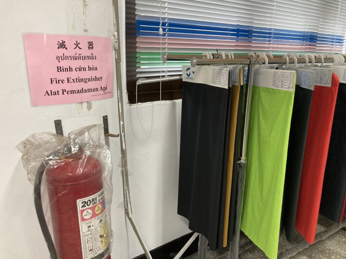 A fire extinguisher mounted on a wall next to multilingual safety instructions and a rack of colorful fabric samples.