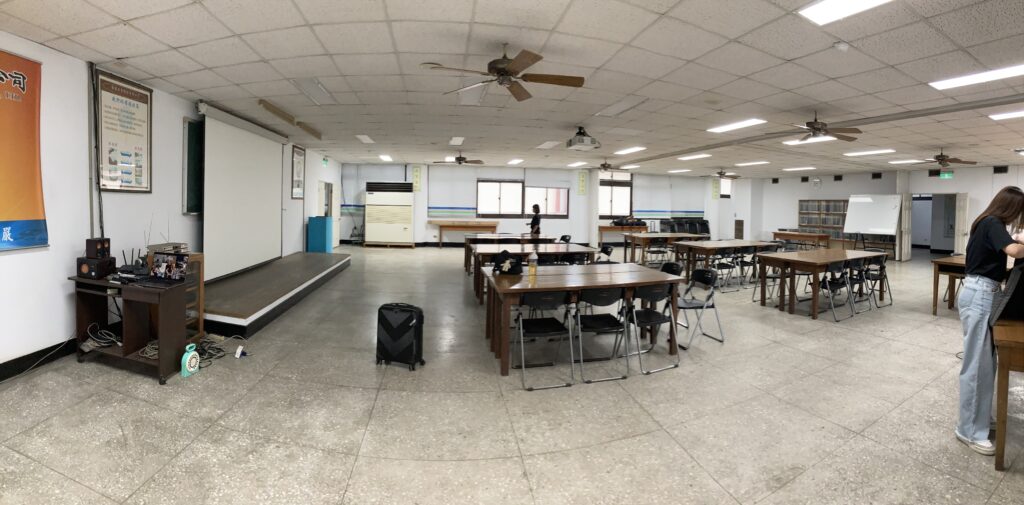 A spacious factory meeting room with rows of wooden tables, chairs, a small stage, and ceiling fans.